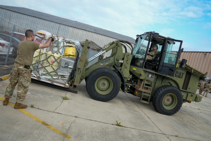 Photo shows large equipment carrying pallet being driven by woman in uniform while being directed by man in uniform