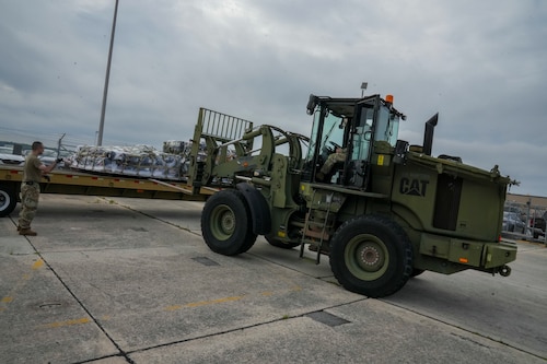 Photos shows large equipment putting pallet onto truck being directed by man in uniform