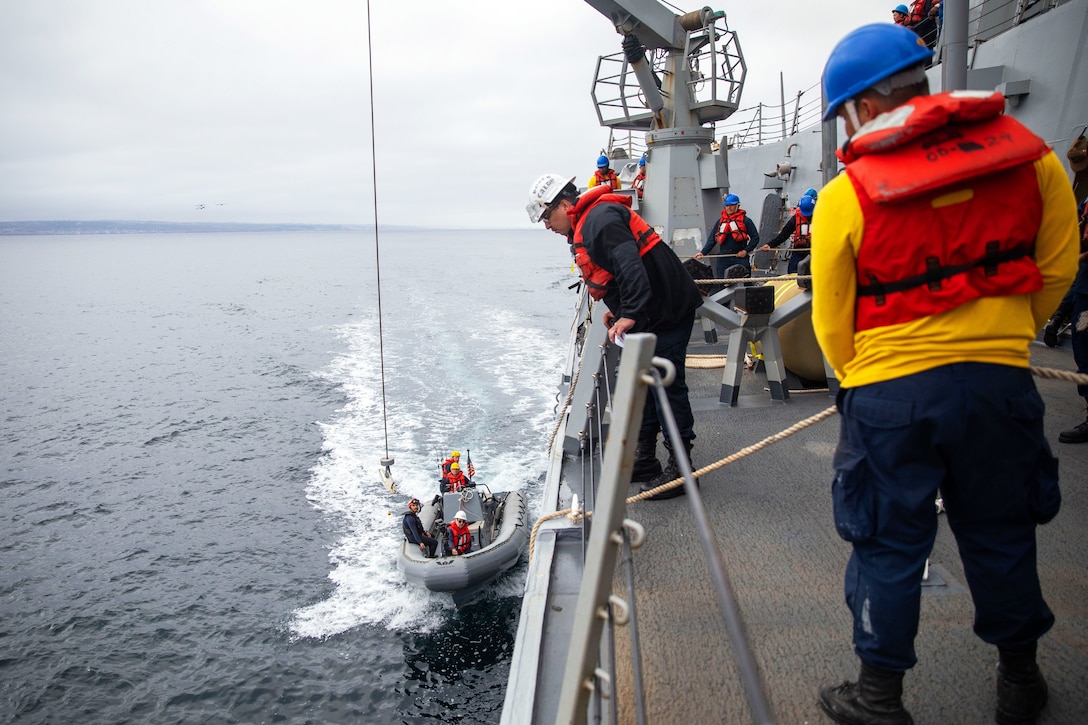 A small inflatable boat approaches the side of a larger ship at sea with sailors on the deck holding ropes.