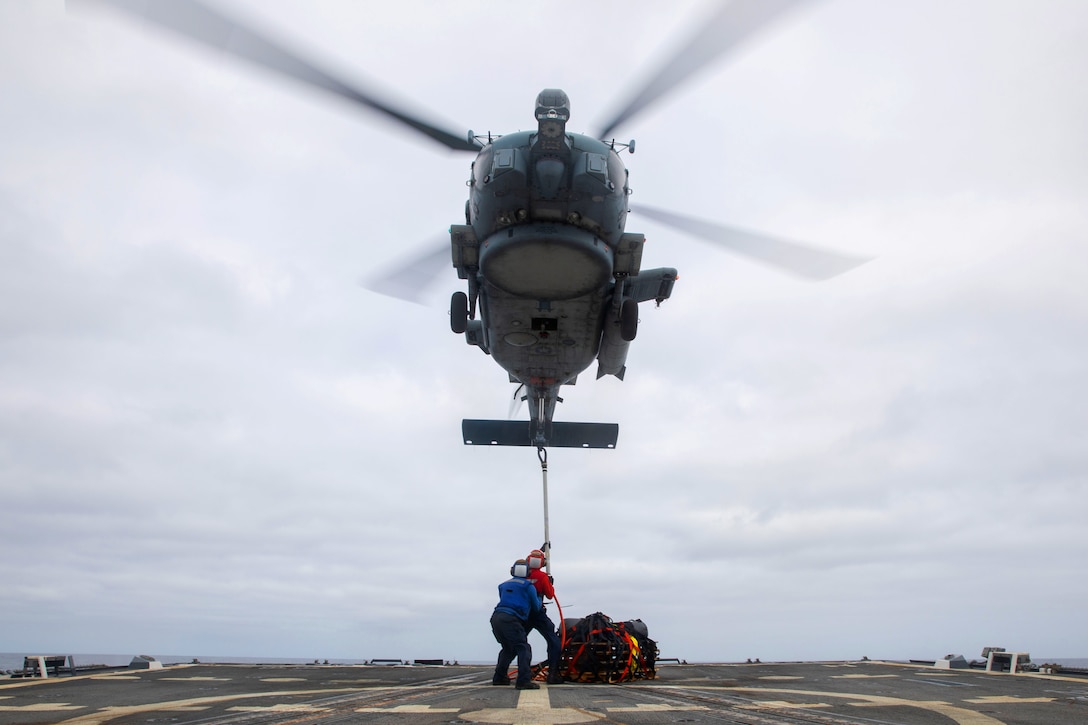 Two sailors on the deck of a ship hook a large cable with cargo attached to a helicopter hovering above them.