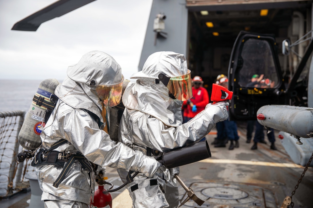 A sailor in firefighting gear holds an extinguisher as another points a thermal device toward the side of a helicopter on the deck of a ship.