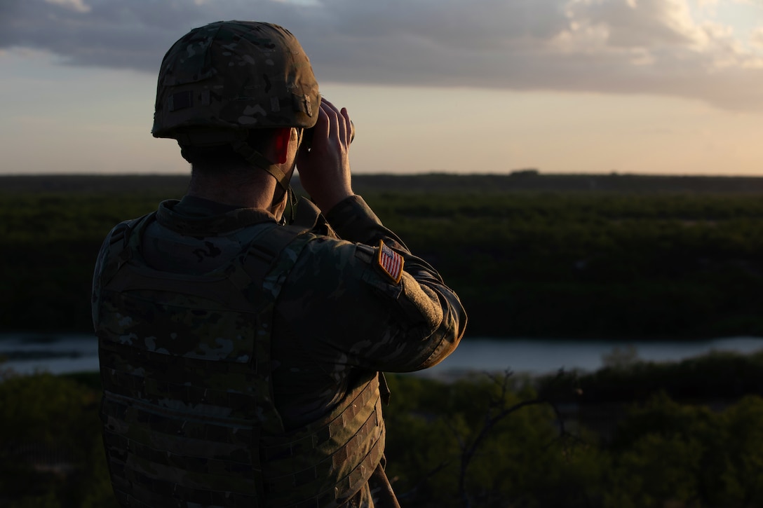 A soldier wearing combat gear and using binoculars looks toward a river with heavy green vegetation on each side.