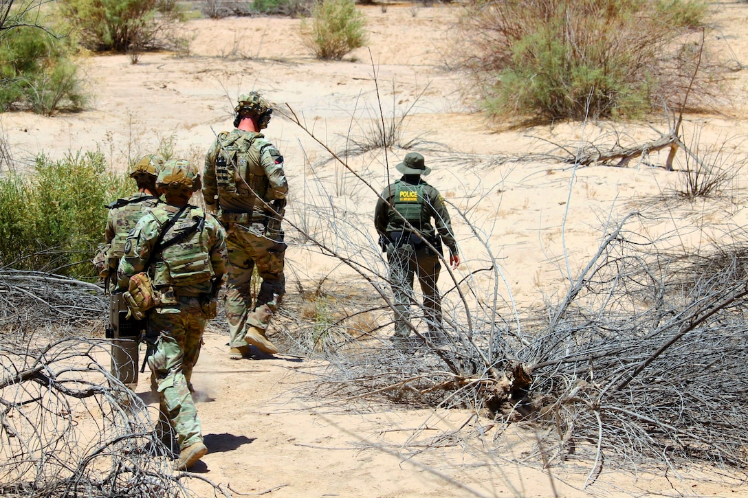 Three soldiers in combat gear walk behind a Border Patrol agent in desert-like conditions on a sunny day.