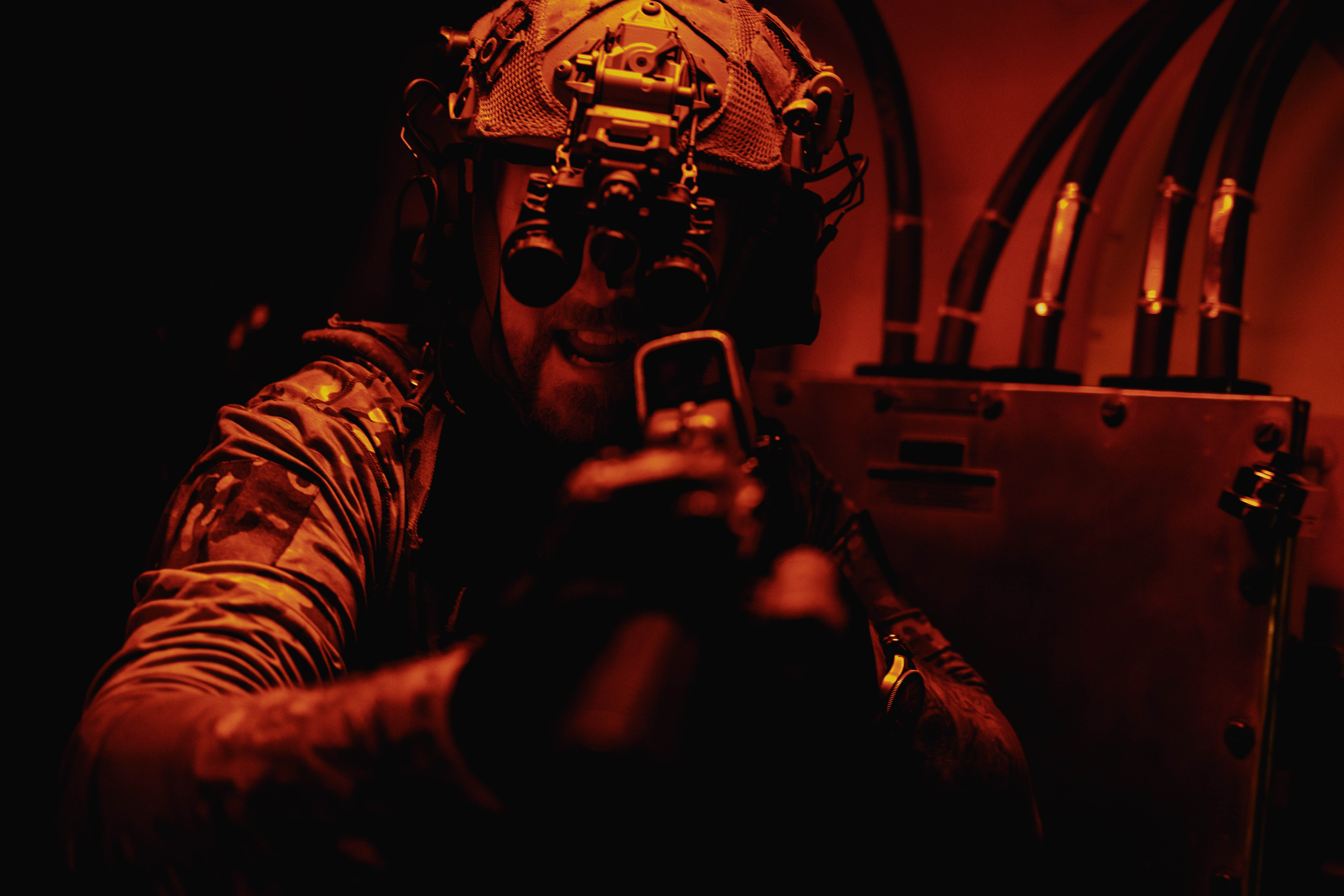 A sailor in tactical gear aims a gun while under a red light.