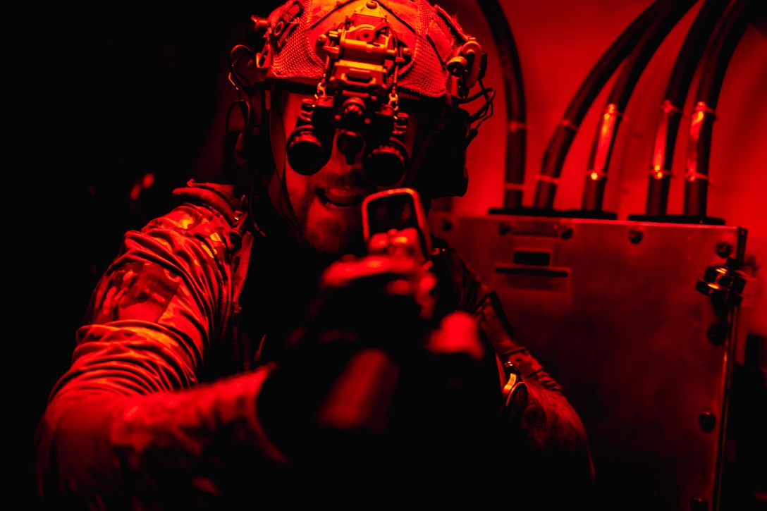 A sailor in tactical gear aims a gun while under a red light.