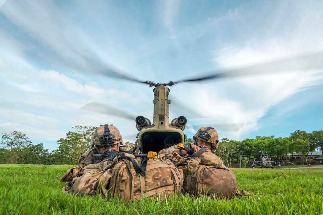Two soldiers aim weapons while laying in a field in front of a helicopter on a sunny day.