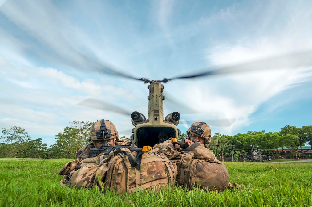 Two soldiers aim weapons while laying in a field in front of a helicopter on a sunny day.