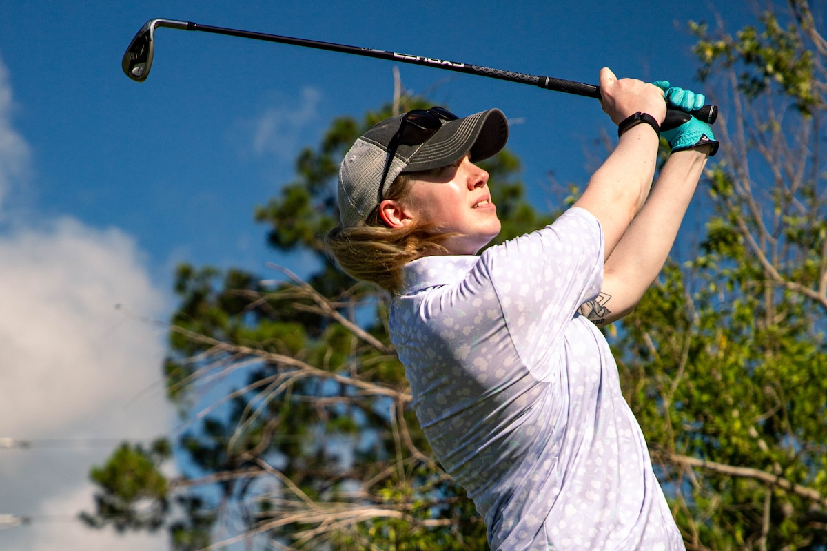 A soldier swings a golf club in front of a tree on a sunny day.