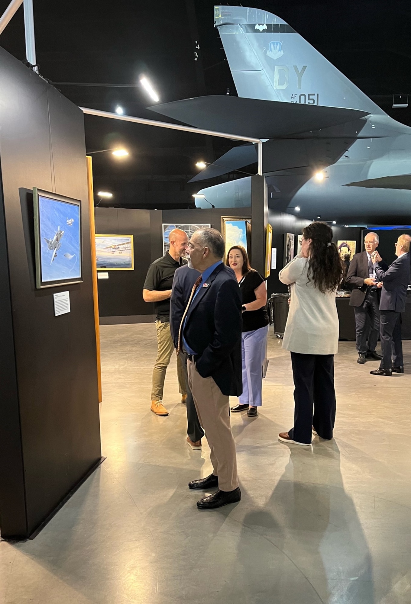 a group of tall black panels showcase artwork with large aircraft in the background and a number of people talking and looking at the artwork in the foreground