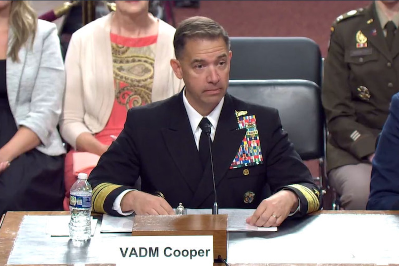 A man wearing a dress military uniform sits at a table in front of a microphone. In front of him is a sign that reads, “VADM Cooper,” and behind him are people wearing business attire and military uniforms.