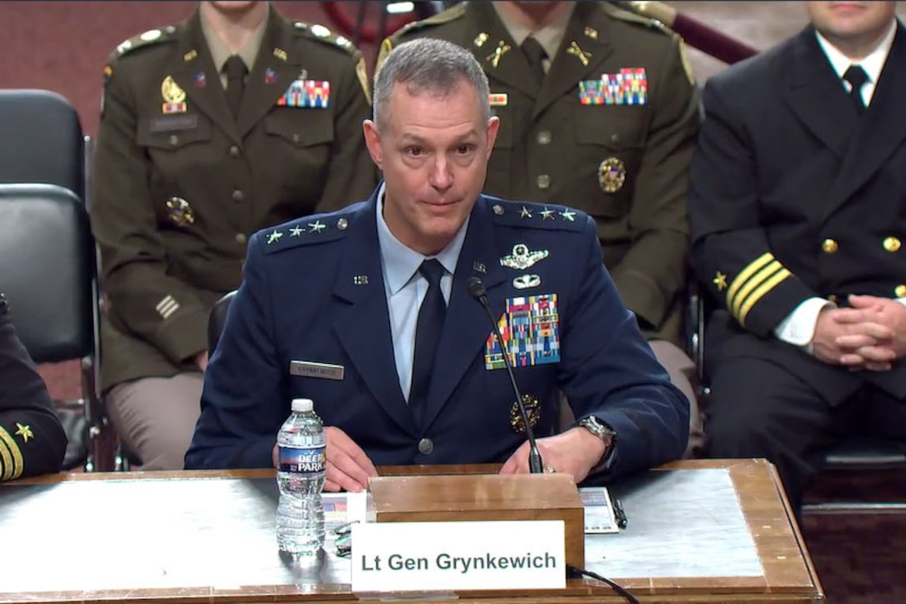 A man wearing a dress military uniform sits at a table in front of a microphone. In front of him is a sign that reads, “Lt Gen Grynkewich,” and behind him are people wearing military uniforms.
