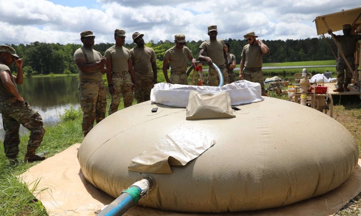 Seven Soldiers stand outside near a lake. One is holding a blue hose that pulls water from the lake. The hose is pushing water into a large tan water holder that looks like a giant bag.