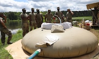 Seven Soldiers stand outside near a lake. One is holding a blue hose that pulls water from the lake. The hose is pushing water into a large tan water holder that looks like a giant bag.