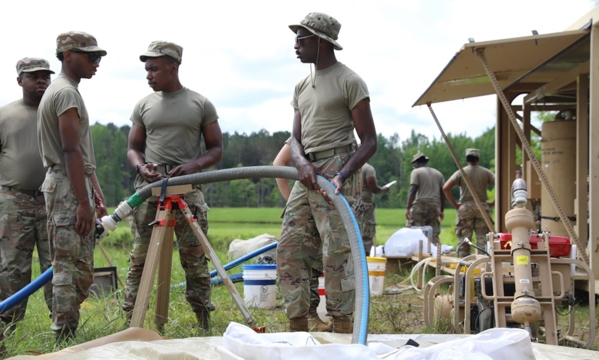 Four Soldiers stand outside holding a blue hose that pulls water from a lake into a holding tank