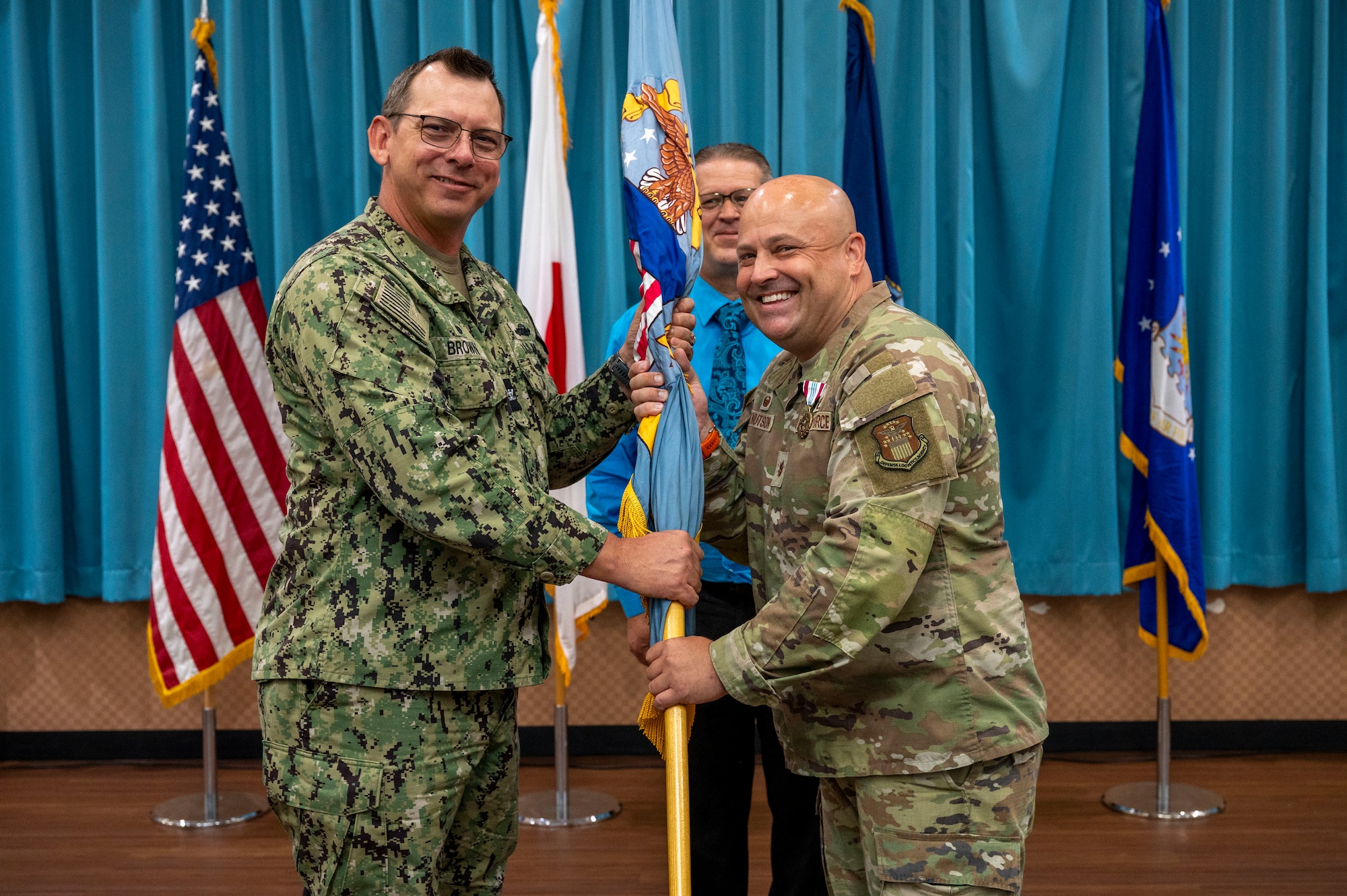 U.S. Navy Capt. Patrick Brown, Defense Logistics Agency Indo-Pacific commander, left, receives the guidon from U.S. Air Force Maj. Kevin Knutson, DLA Energy Okinawa outgoing commander, right, during  a change of command ceremony at Kadena Air Base, Japan, June 18, 2025.