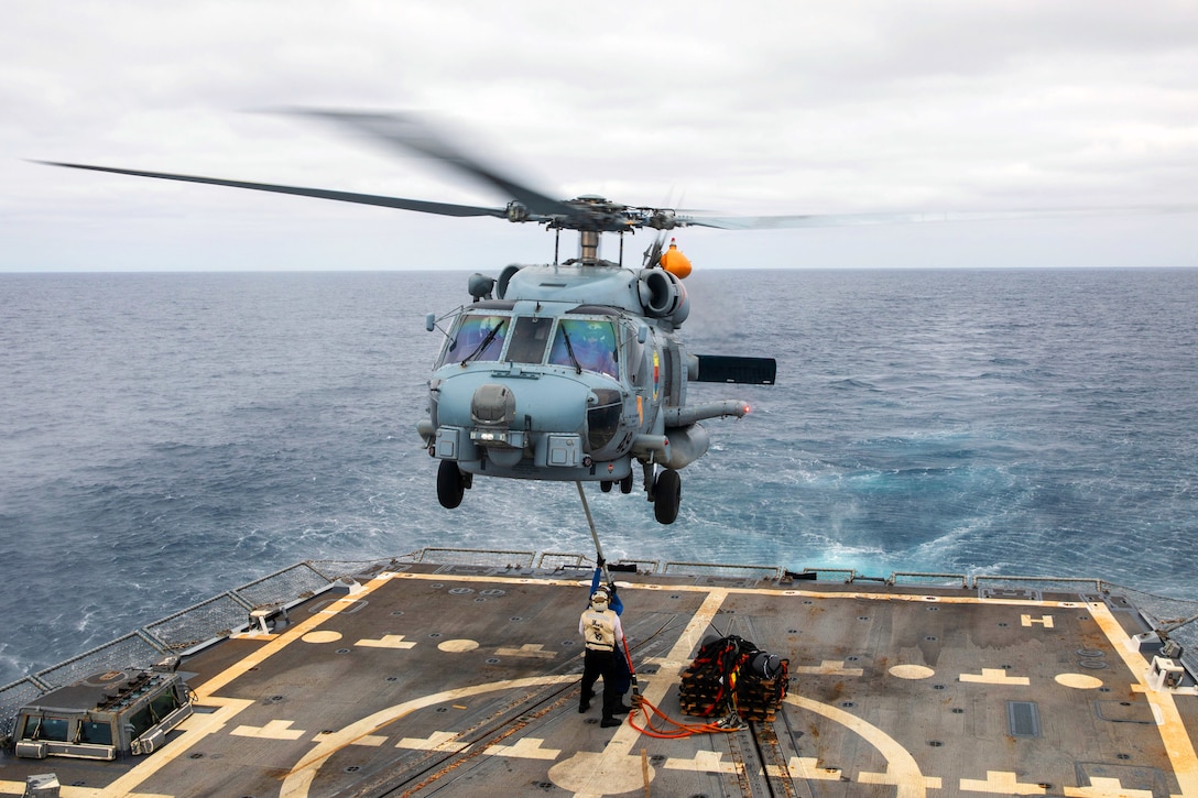 Two sailors hook cargo to the underside of a helicopter hovering above the deck of a ship at sea on a cloudy day.