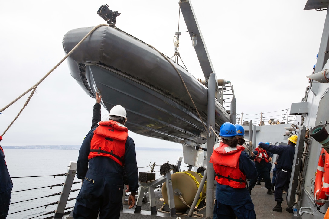 Sailors wearing helmets and life jackets use ropes and cables to hoist a small boat onto a larger ship.