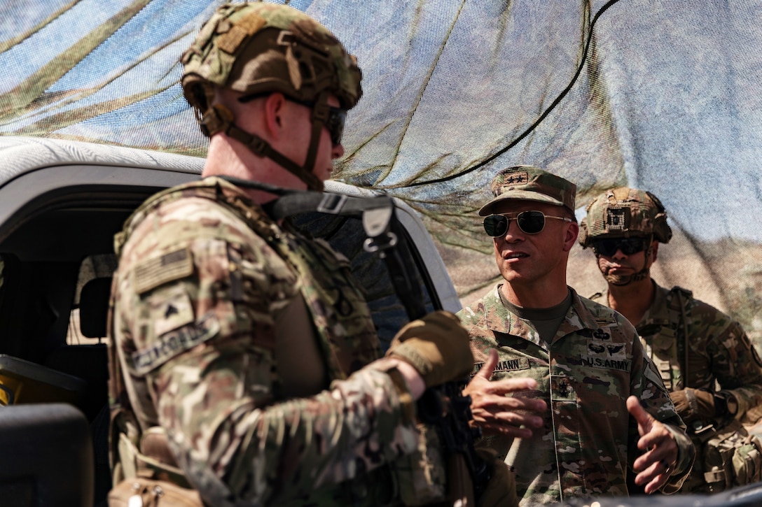 Two soldiers standing next to a vehicle and under camouflage netting talk as another looks on in the background.