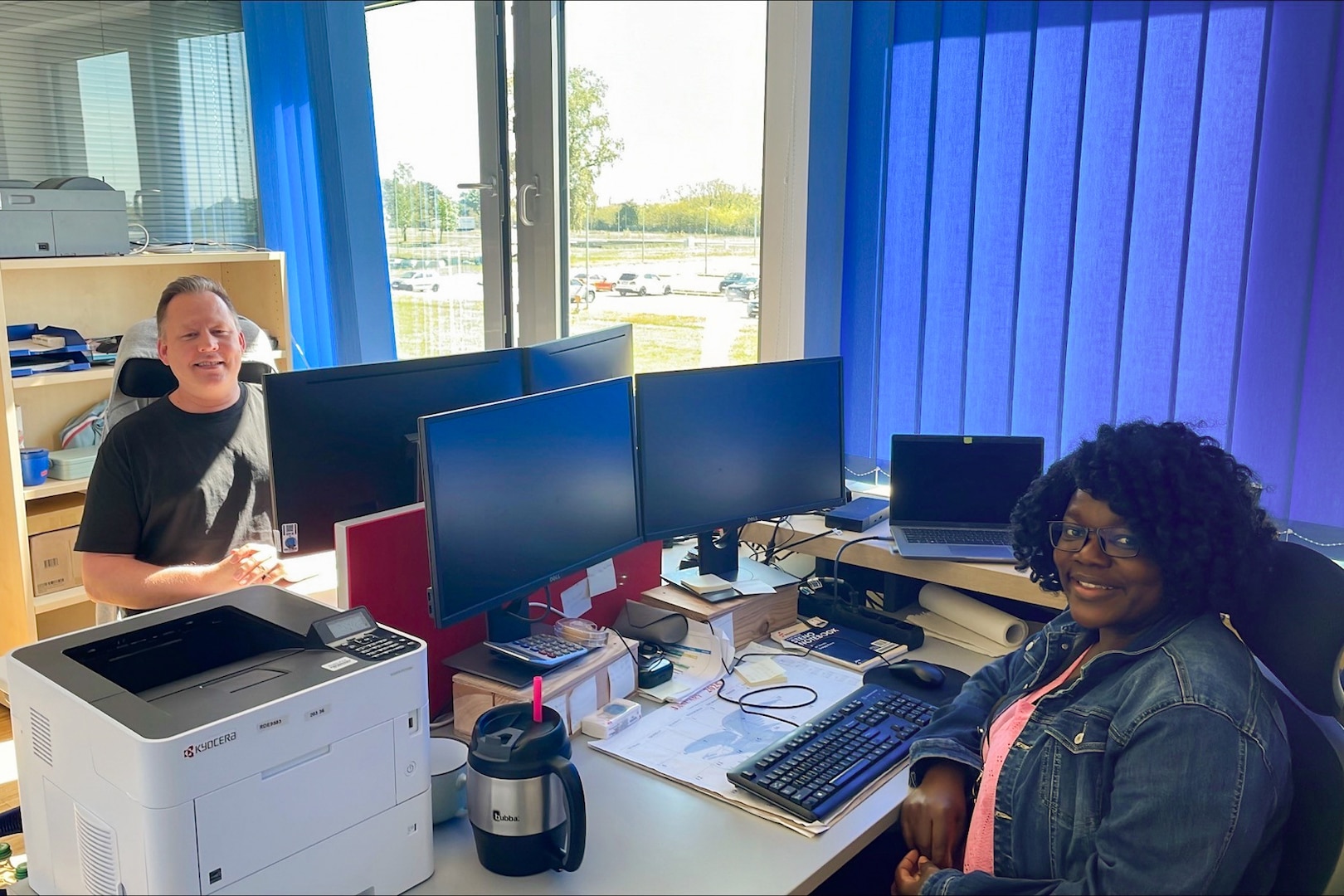 A man and a woman sit behind their desks that face one another. They are both looking at the camera.