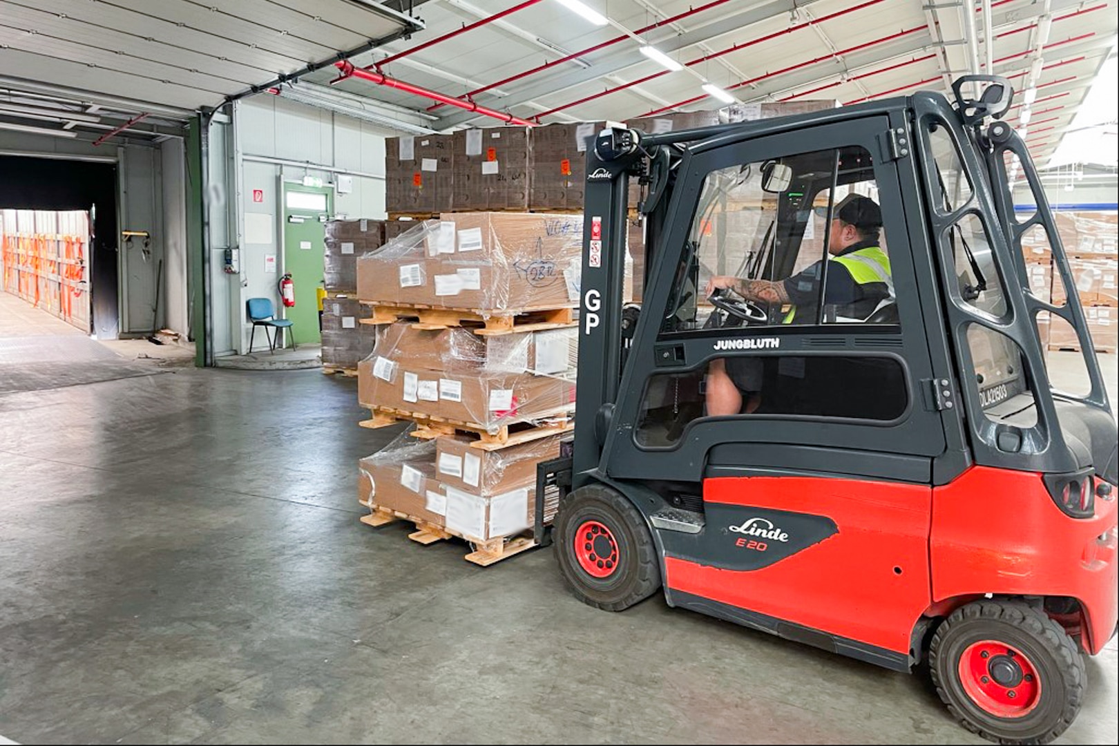 A man driving a forklift in a warehouse uses the forklift to stack pallets of brown boxes.