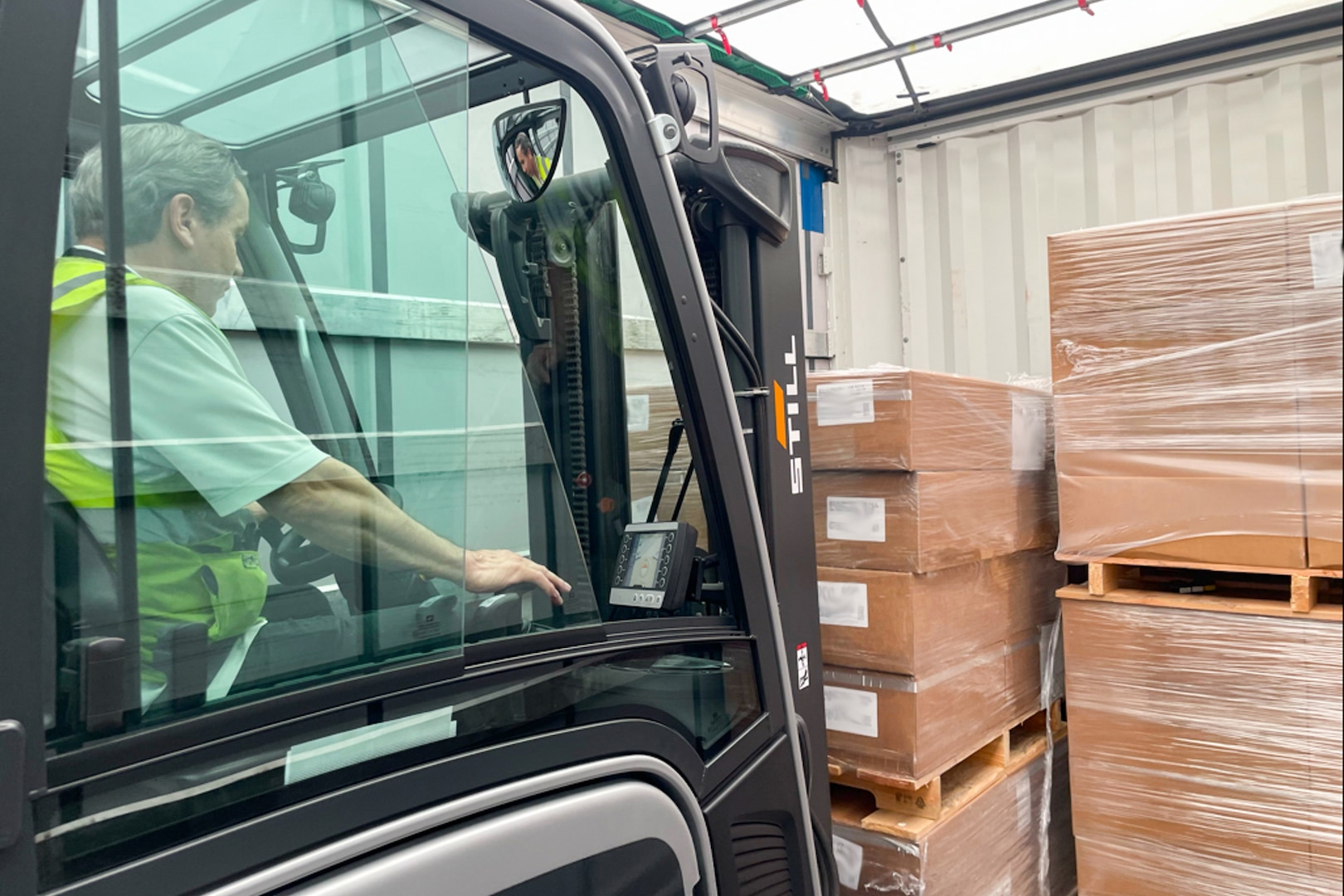 A man driving a forklift in a warehouse uses the forklift to stack pallets of brown boxes.