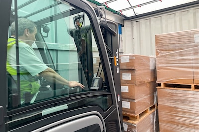 A man driving a forklift in a warehouse uses the forklift to stack pallets of brown boxes.