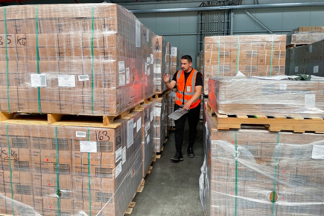 A man wearing an orange safety vest walks between stacked pallets of brown boxes in a warehouse.