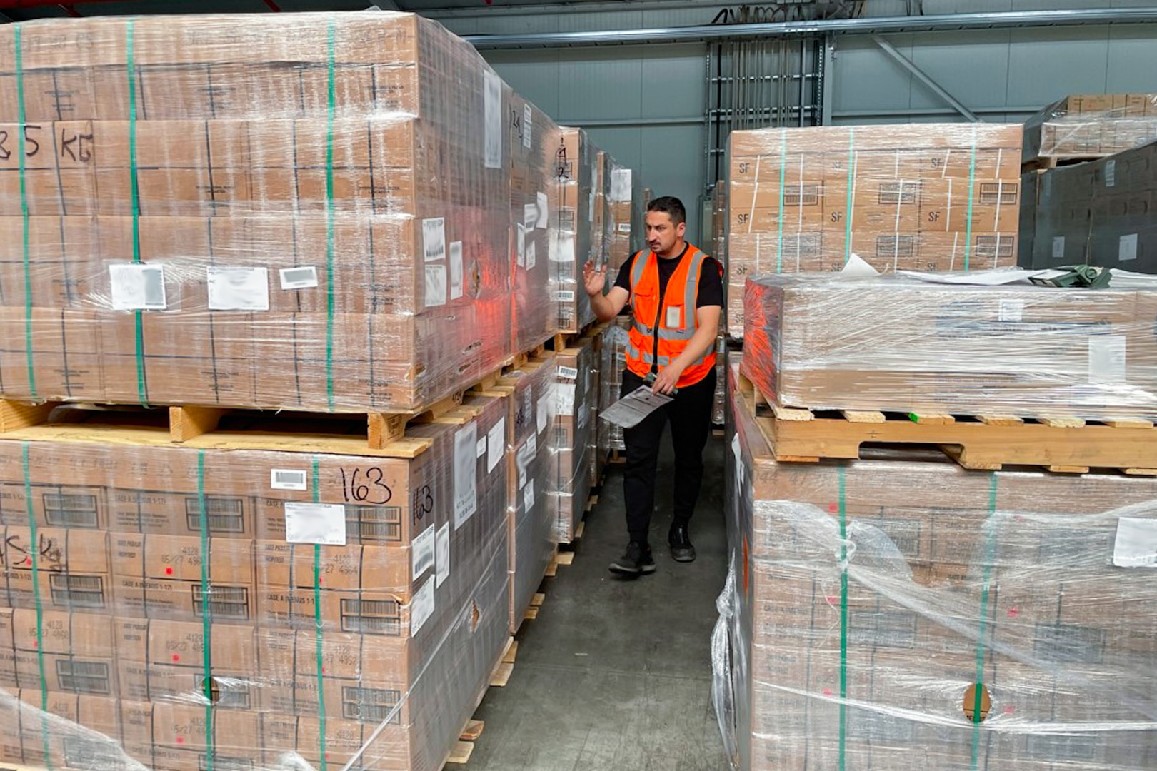 A man wearing an orange safety vest walks between stacked pallets of brown boxes in a warehouse.