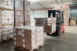 A man driving a forklift in a warehouse uses the forklift to stack pallets of brown boxes.