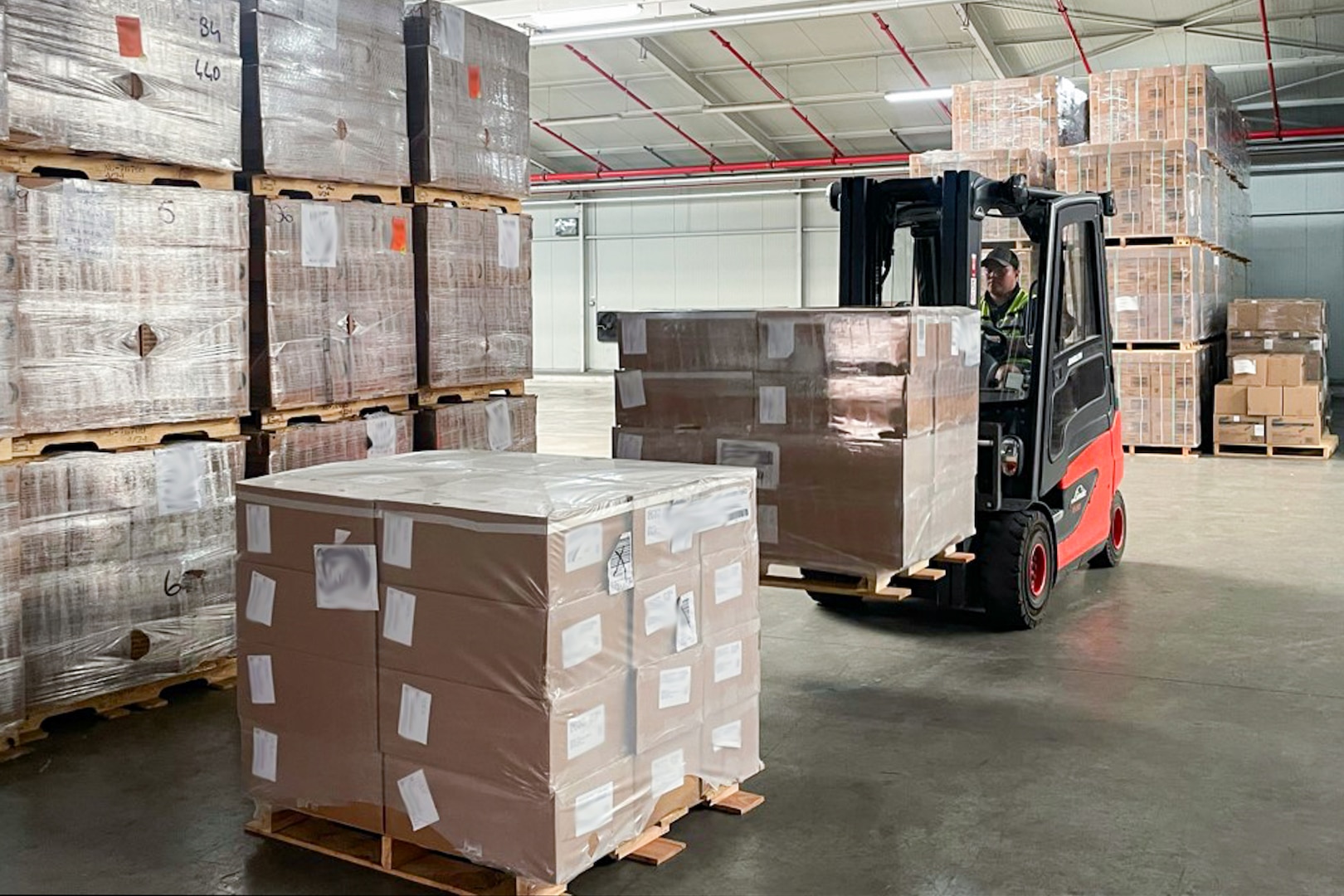 A man driving a forklift in a warehouse uses the forklift to stack pallets of brown boxes.