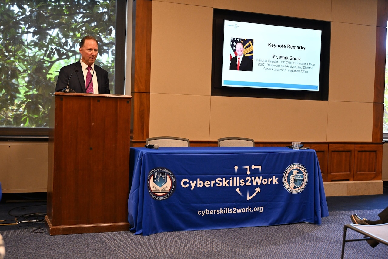 A man in business attire is standing in a room behind a podium, speaking into a microphone. A table with a cloth that reads “CyberSkills2Work” is next to him.