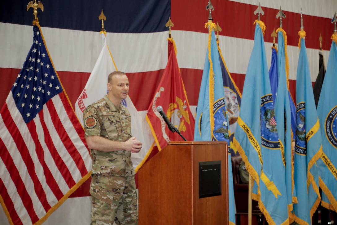 A man wears a camouflage outfit while standing at a podium in front of flags