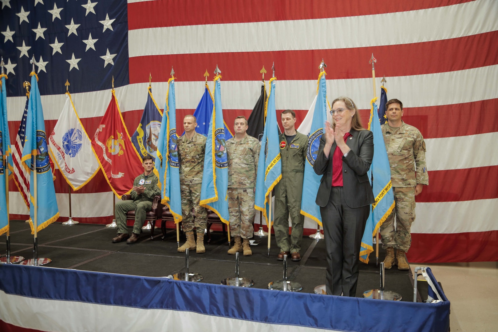 A female stands at the podium clapping while others stand at attention in front of flags