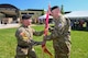 Two people wearing military uniforms participate in an outdoor change of command ceremony. One passes a flag to another.