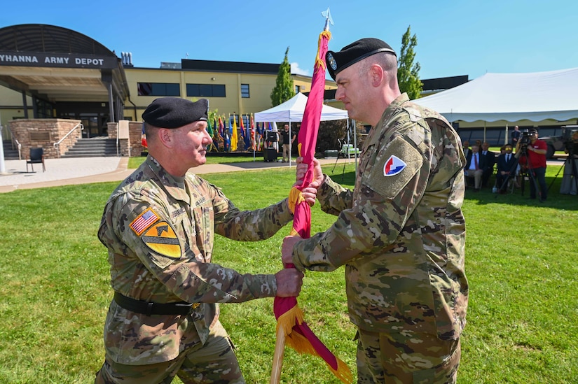 Two people wearing military uniforms participate in an outdoor change of command ceremony. One passes a flag to another.
