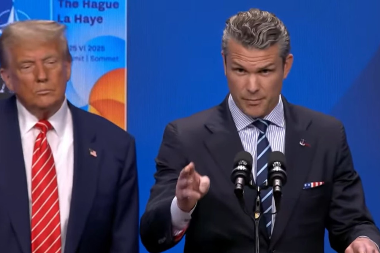 A man in a suit and tie speaks in front of a lectern while another man in in a suit beside him looks on.