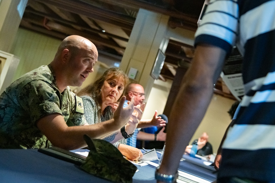 A man wearing a camouflage military uniform and woman sit at a table looking across the table at another person.