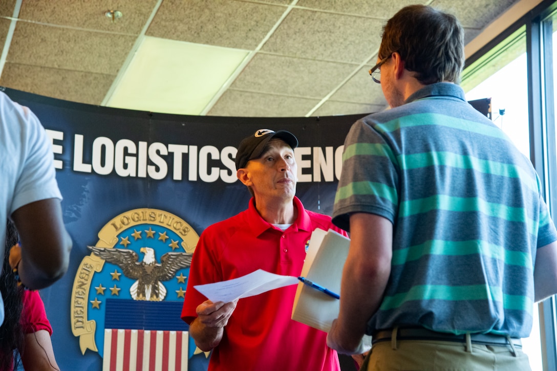 A man in a red shirt stands facing another man holding papers.