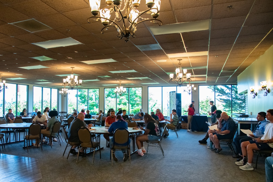 A large group of people sit at tables in a large ballroom event space lined with floor-to-ceiling windows.