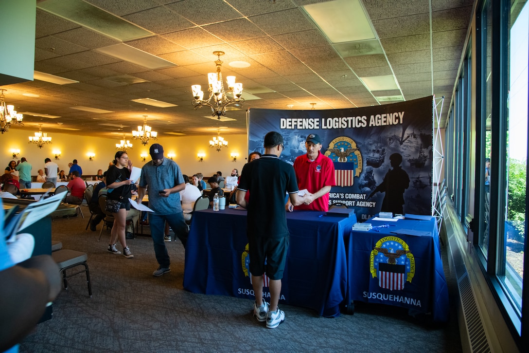 A man stands behind a table in front of a backdrop with the words 'Defense Logistics Agency' on it, helping a person on the other side of the table. There are other people in the background.