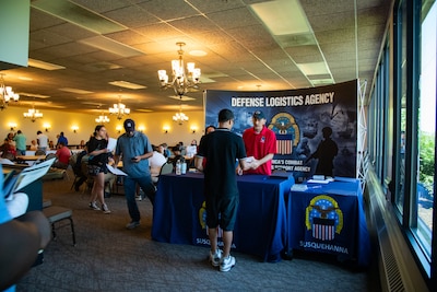 A man stands behind a table in front of a backdrop with the words 'Defense Logistics Agency' on it, helping a person on the other side of the table. There are other people in the background.
