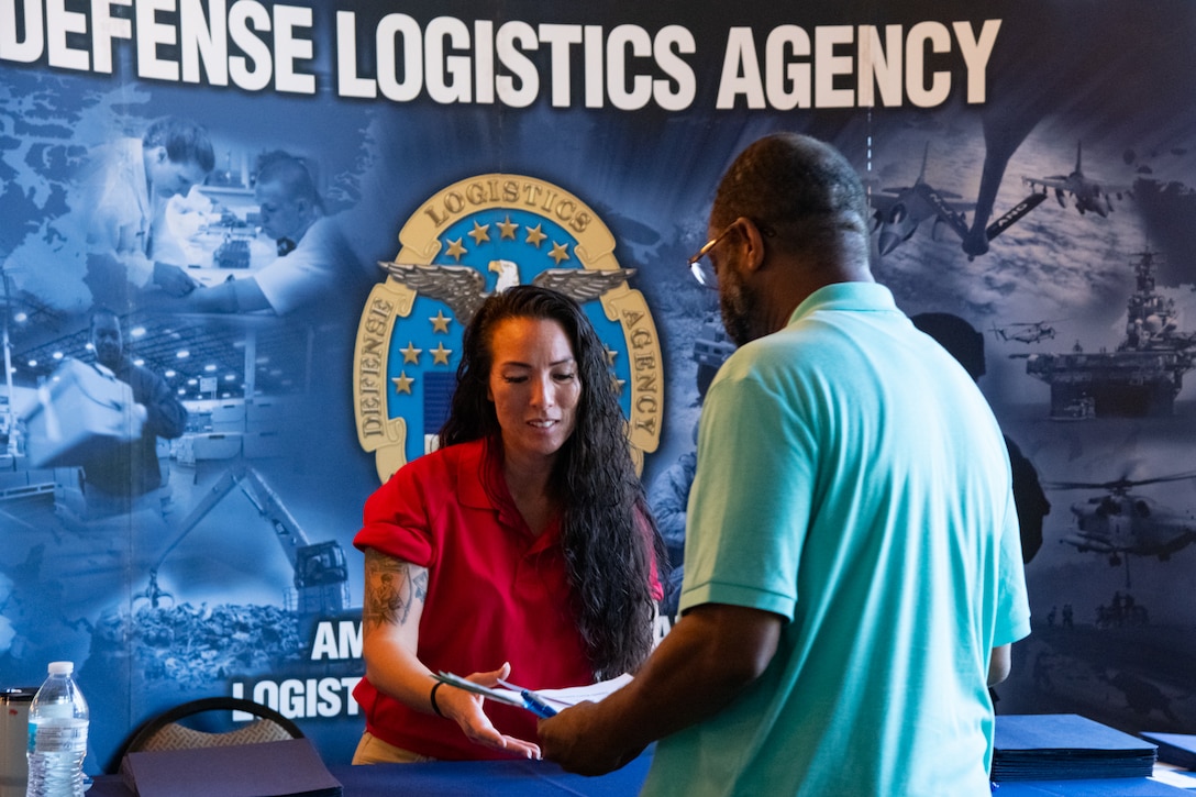 A woman in a red shirt takes papers from a man. There is a backdrop behind her that says 'Defense Logistics Agency'.