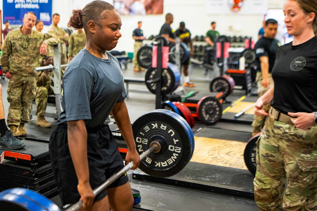 A soldier lifts a barbell with four weights on it as other soldiers look on inside a gym.