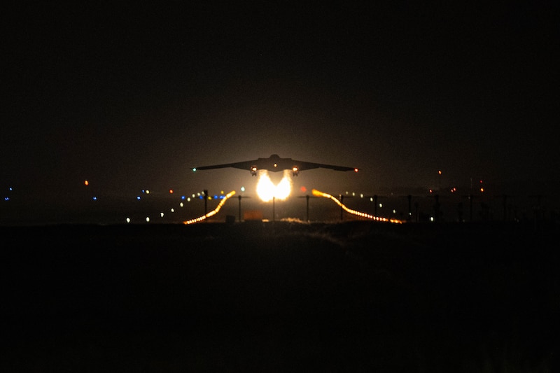 A B-2 bomber aircraft lifts off from a dark runway, trailing two fire-like contrails with lights in the background.