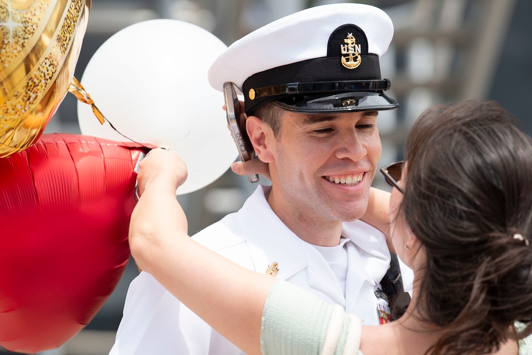 A smiling sailor in a white uniform hugs a person holding balloons and carrying a bag on a sunny day.