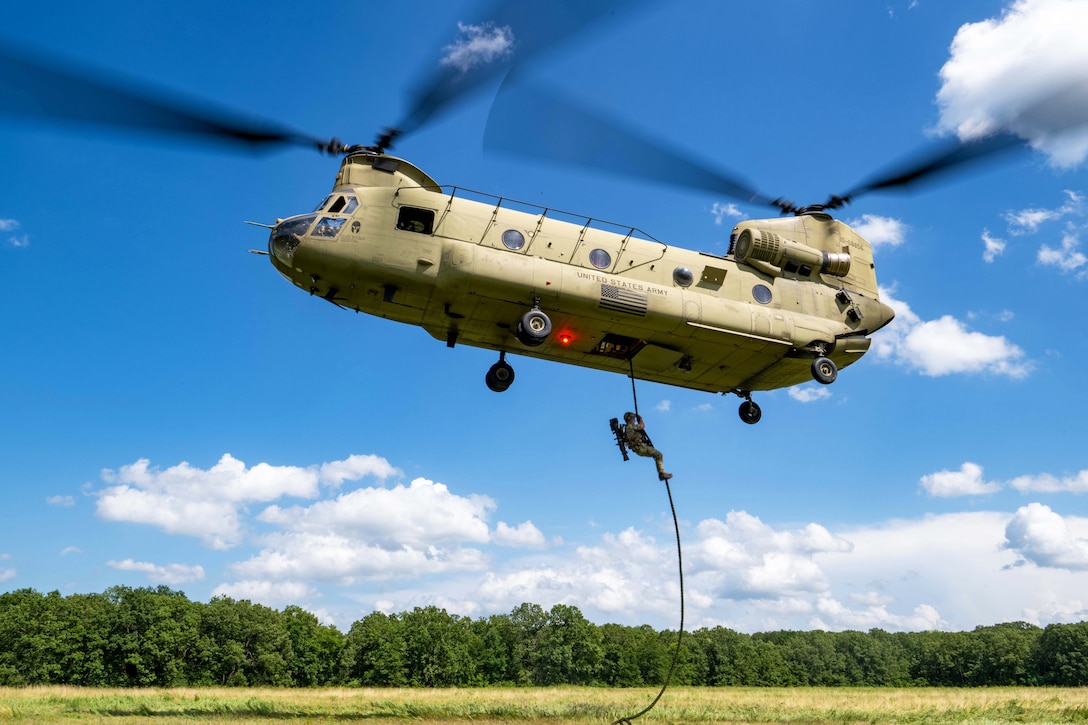 A Navy SEAL uses a rope attached to a hovering helicopter to descend to a grassy field on a sunny day.