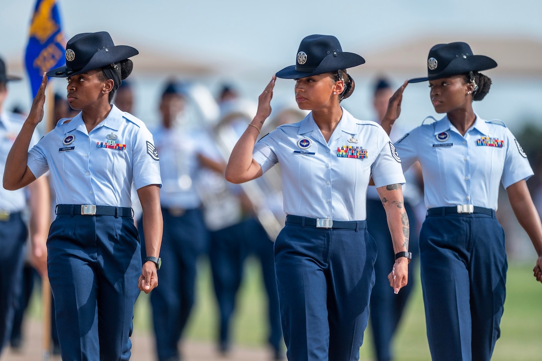Three airmen turn their heads and salute while marching outdoors, with other airmen in the background.