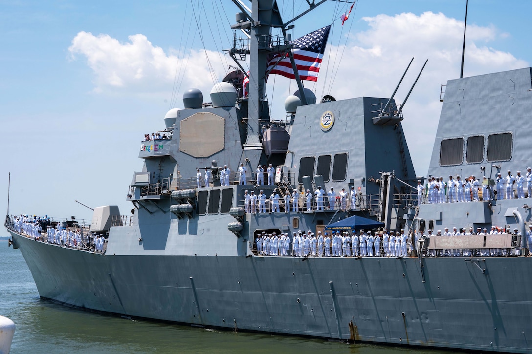 Sailors stand at attention on the deck of a ship flying a large American flag as it leaves port on a sunny day.