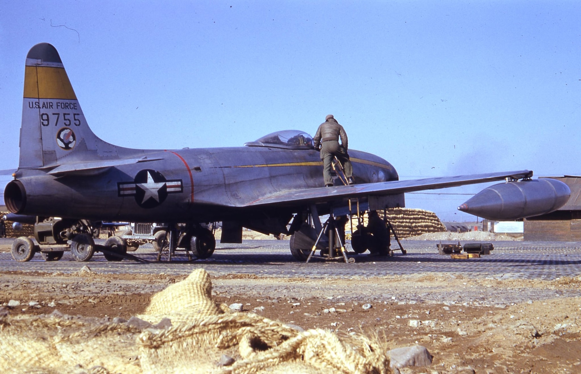 One of the 41st FIS F-80C Shooting Star jet fighters, serial number 49-755, undergoes maintenance at a forward airfield during the Korean War.  Note the squadron emblem, the Flying Buzz Saw in the Clouds, on the tail, as well as the pierced steel planks the aircraft rests on and the sandbag walls of aircraft revetments beyond.  The aircraft was later assigned to the 80th Fighter-Bomber Squadron and was lost in a takeoff accident in Korea during May, 1952.  (Photo courtesy of Mr. Jack Cook)