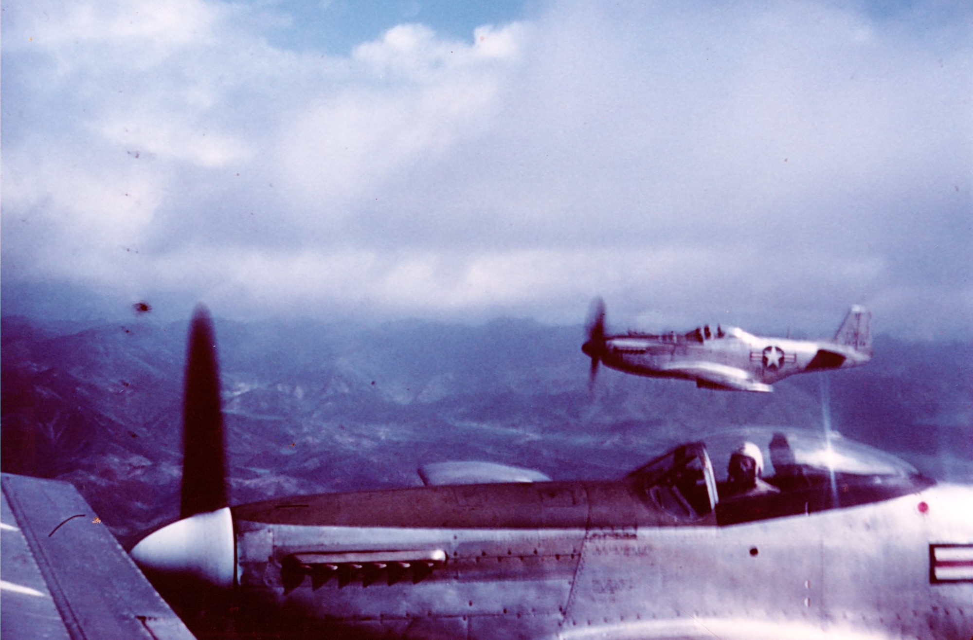 Capt. Ernie Biggs tucks it in tight as he leads Capt. Alex Currie in the background in F-51D Mustangs of the 39th FIS over some rugged countryside in East Asia, perhaps on the return to base from a combat mission in Korea, as Currie’s aircraft is seen in flight without any external ordnance.  (Photo courtesy of Mr. Jack Cook)
